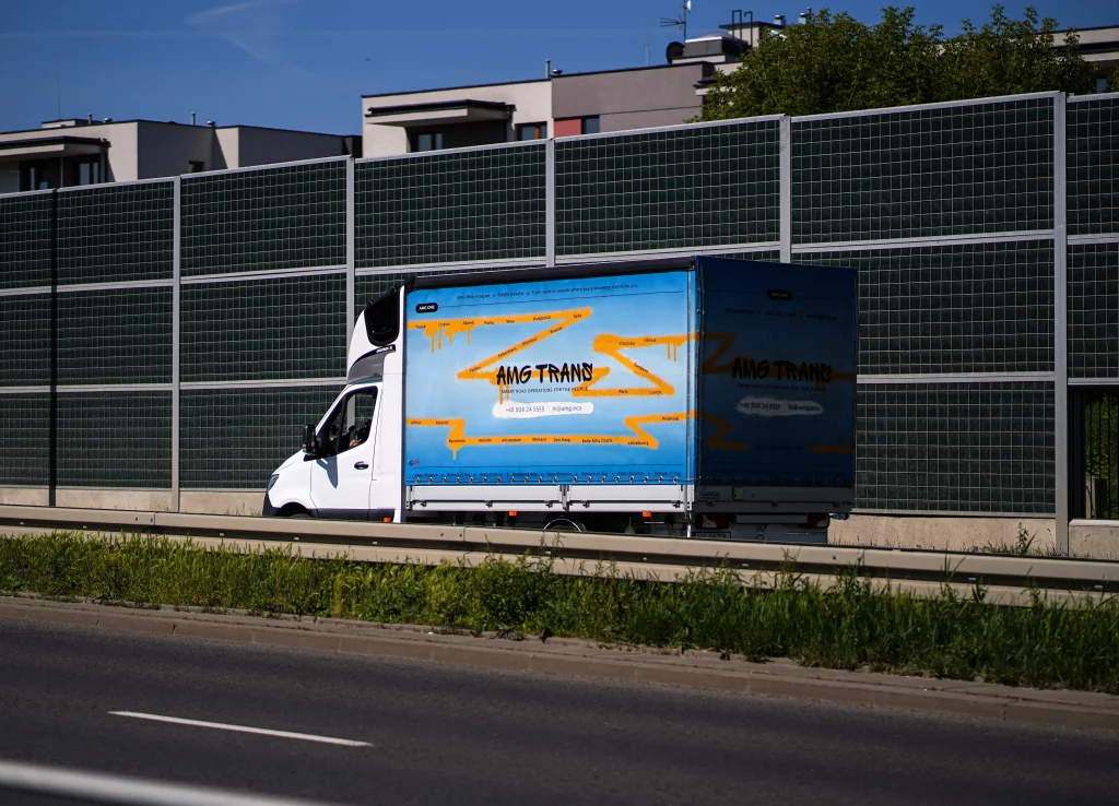 Bus AMG Trans do 3,5T podczas jazdy miejskiej – realizacja dostawy w modelu door-to-door, tło z ekranami dźwiękochłonnymi i zabudową mieszkaniową.