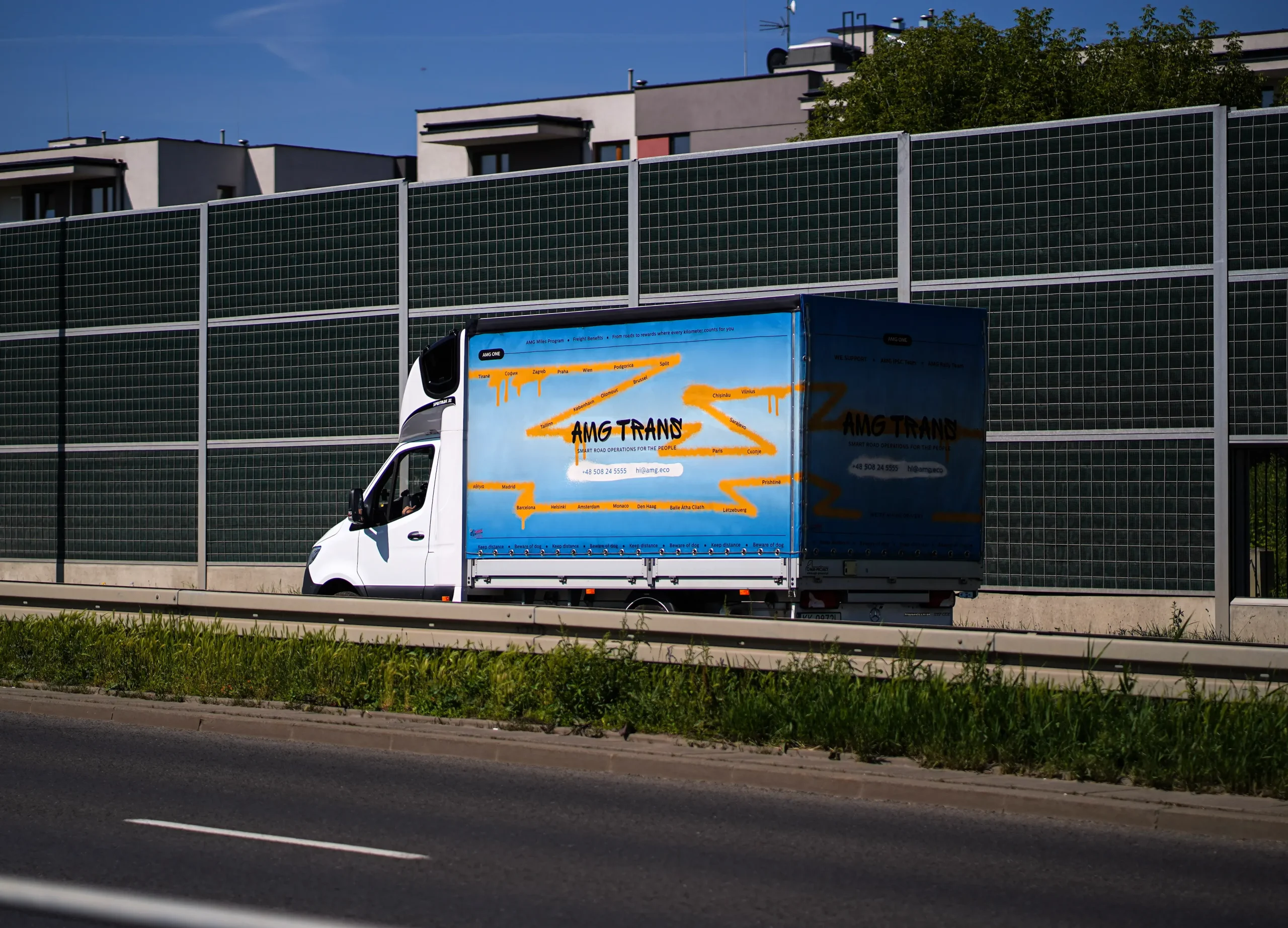 Bus AMG Trans do 3,5T podczas jazdy miejskiej – realizacja dostawy w modelu door-to-door, tło z ekranami dźwiękochłonnymi i zabudową mieszkaniową.