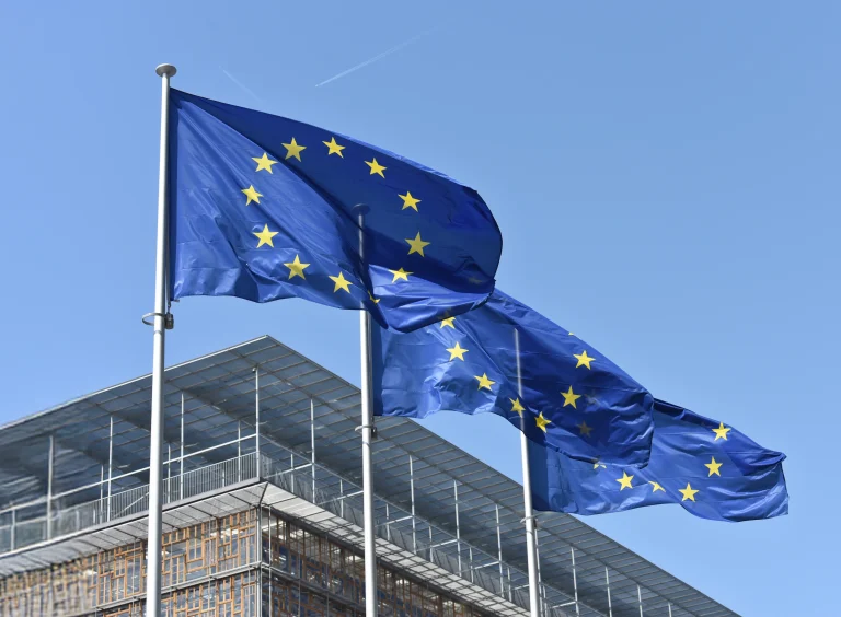 Trzy flagi Unii Europejskiej powiewające na masztach przed budynkiem UE na tle nieba